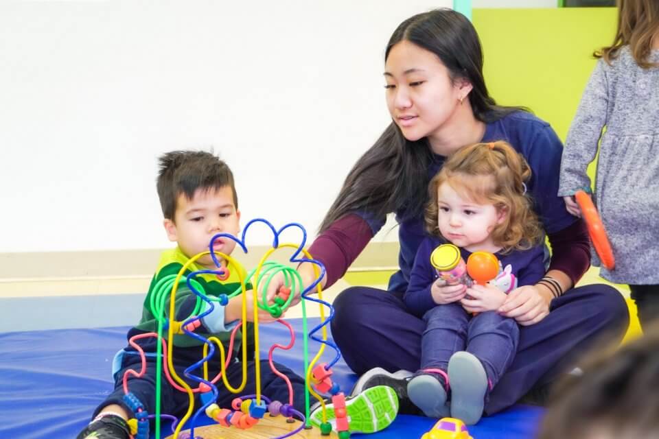 Two children play with colorful toys on a mat while a woman seated beside them observes and assists.