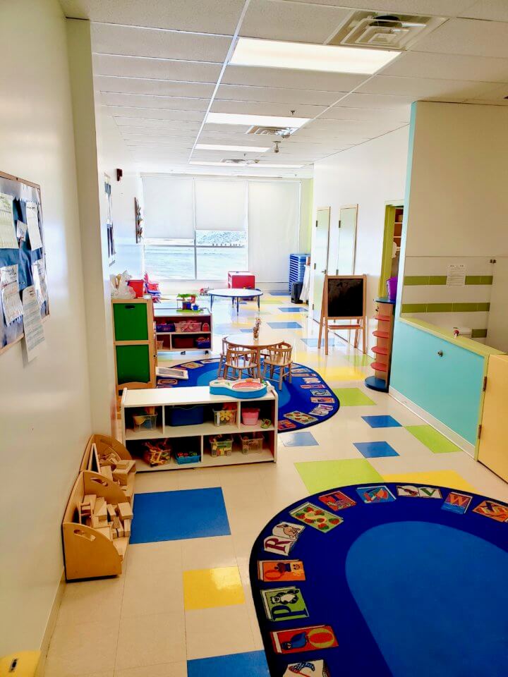 A brightly lit preschool classroom with colorful floor tiles, bookshelves with toys, a round rug with alphabet designs, tables, chairs, and a large window in the background.