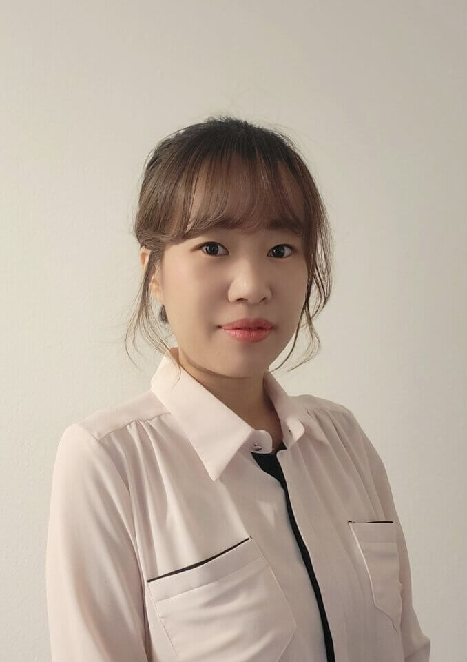 A woman with medium-length brown hair tied back is wearing a light-colored blouse and standing against a plain white background.