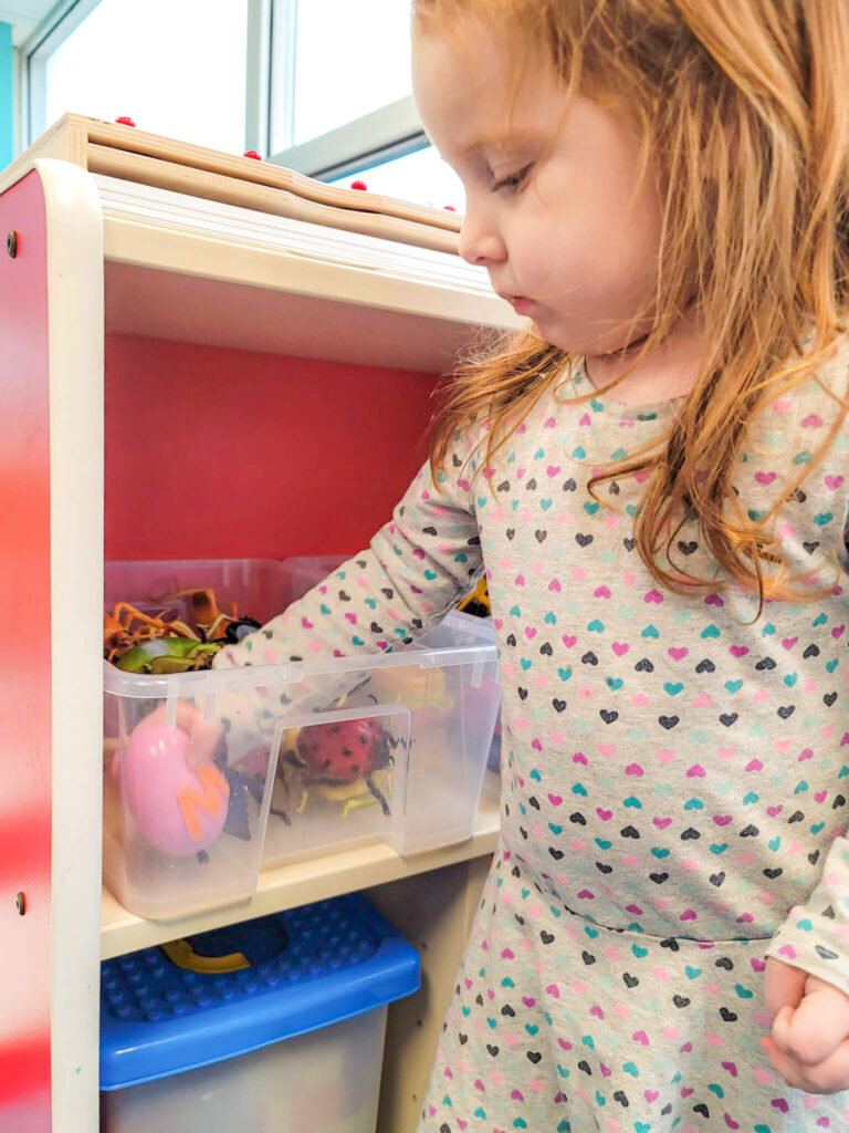 A young girl in a grey dress with heart patterns reaches into a clear plastic bin on a white and red shelf.