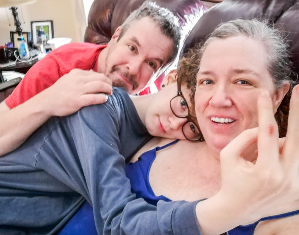 Three people are sitting close together on a couch. One person in glasses is in the center