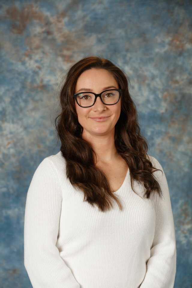 A person with long, wavy brown hair and glasses wearing a white sweater stands in front of a blue and brown mottled background.