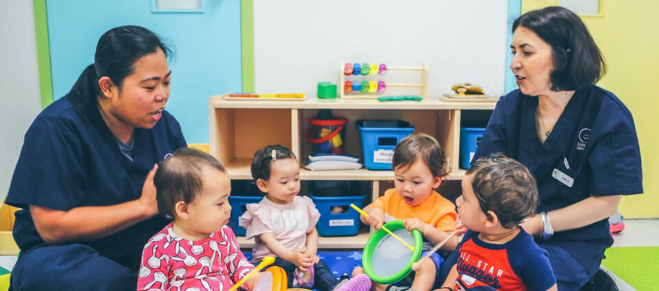 Two caregivers sit with five young children on the floor in a brightly lit room, engaging with toys and musical instruments. Shelving with toys and supplies is visible in the background.