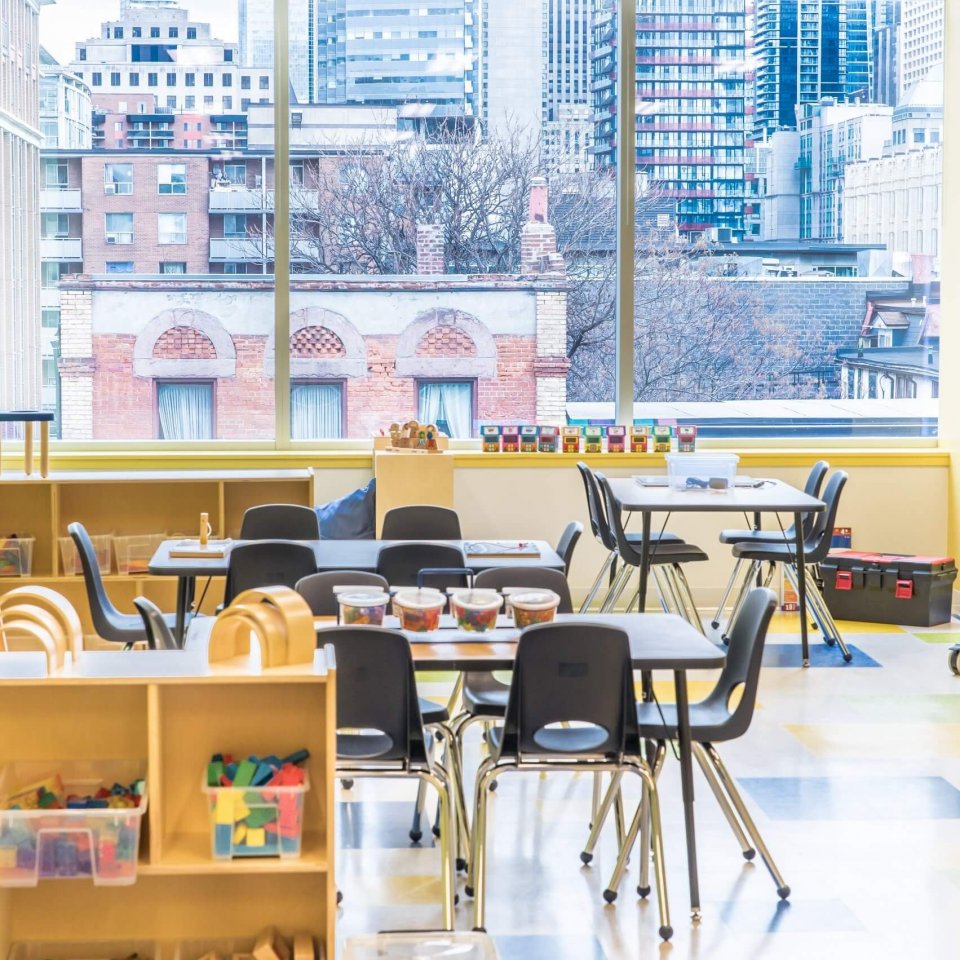 A bright classroom with light-colored furniture, several tables and black chairs, and shelves filled with educational materials. Large windows reveal a cityscape with buildings in the background.