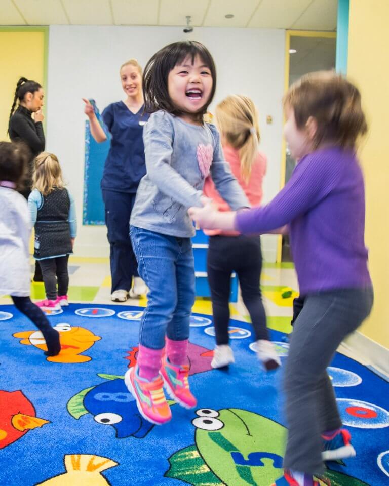 Young children play and jump on a colorful fish-themed rug in a classroom while a smiling adult supervises.