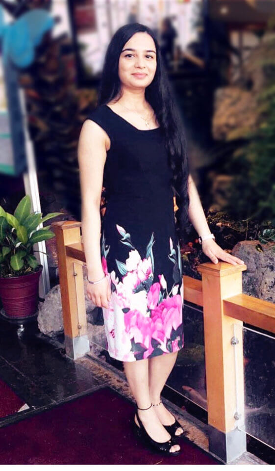 A woman with long, dark hair is standing next to a wooden railing, wearing a black dress with a floral pattern near the bottom and black high heels. There are plants and rocks in the background.