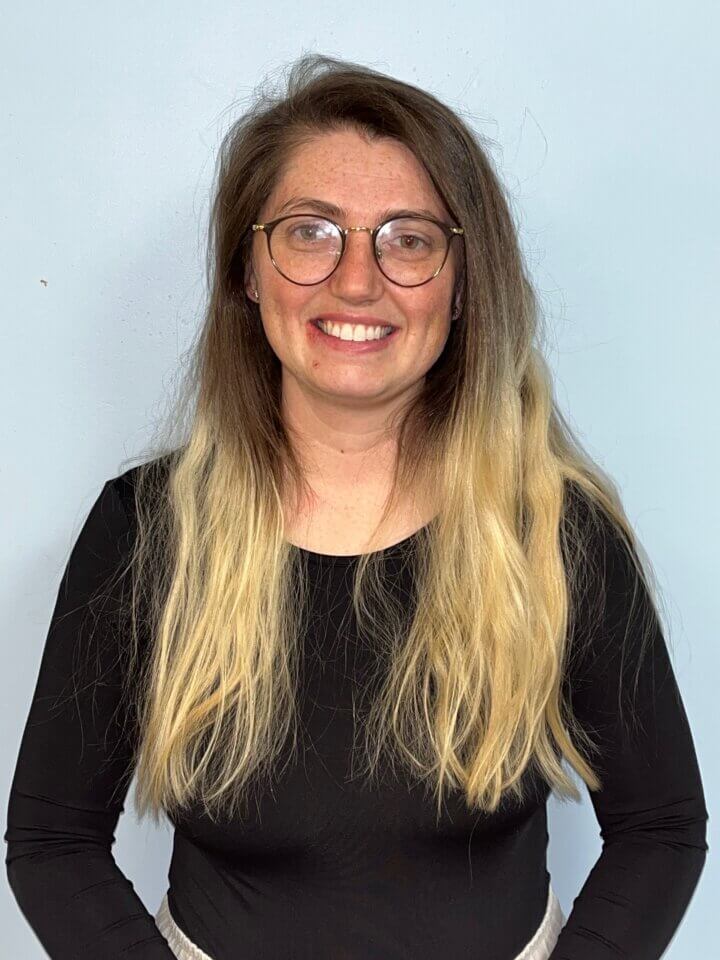 A woman with long, wavy, light brown and blonde hair wearing glasses and a black top stands against a light blue background, smiling at the camera.