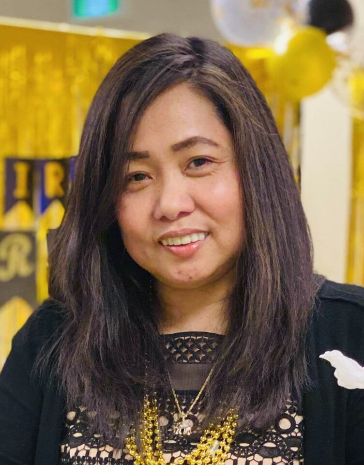 A woman with long dark hair wearing a black top and gold necklace smiles at the camera. The background includes gold decorations.