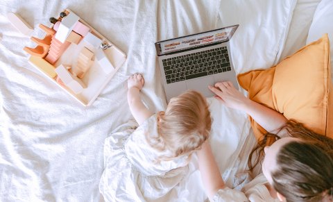 A woman and a child are lying on a bed. The woman is using a laptop, and the child is sitting beside her, looking at the screen. A toy house is placed on the bed next to them.