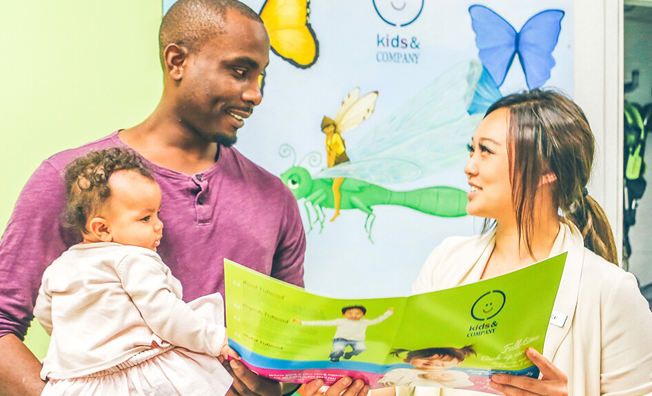 An adult male and female look at a brochure together. The male is holding a baby. A poster with butterflies and the text “kids & COMPANY” is visible in the background.