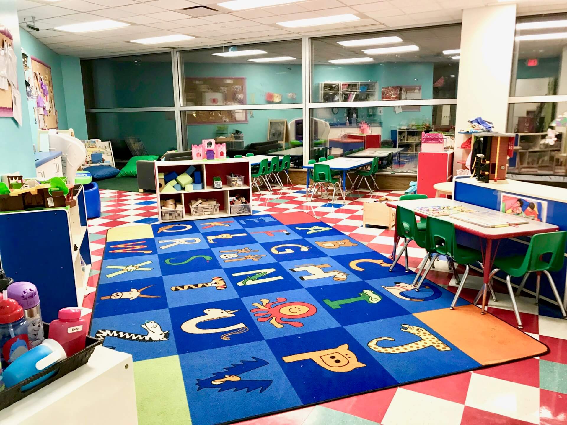 A brightly lit classroom with educational posters, colorful chairs, and an alphabet-themed play mat on the floor. Shelves with toys, books, and learning materials are arranged around the room.