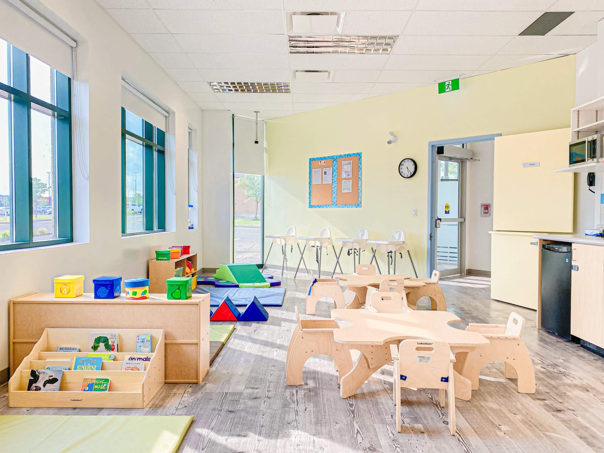A brightly lit classroom with wooden furniture, including small tables and chairs, a bookshelf with picture books, and colorful toys. There are large windows, a clock on the wall, and a green exit sign.
