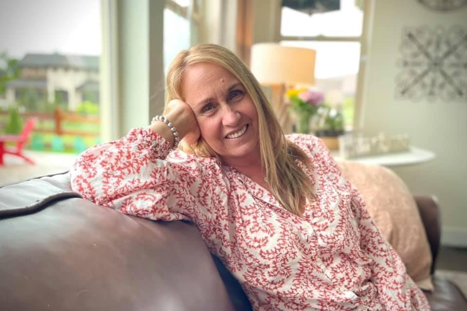 A woman with long blonde hair wearing a red and white patterned blouse is sitting on a couch, leaning her head on her hand and smiling. There's a window and lamp in the background.