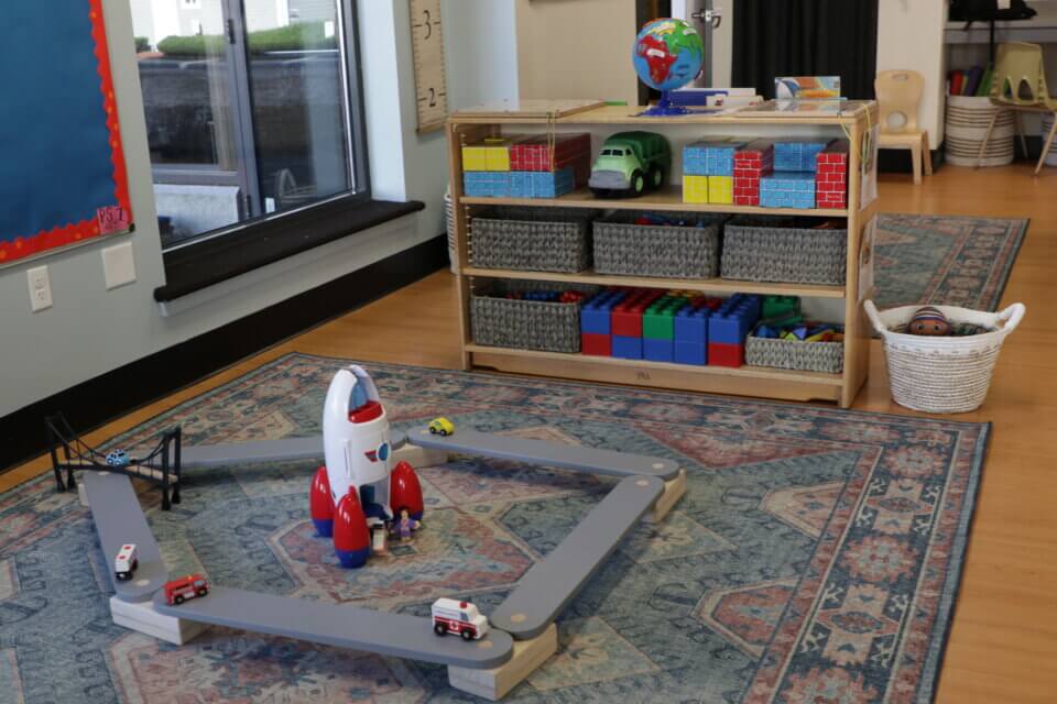 A children's playroom with a rocket-themed toy set on a rug, a shelf with assorted toy bins, a globe on top, and a basket of small toys on the floor.