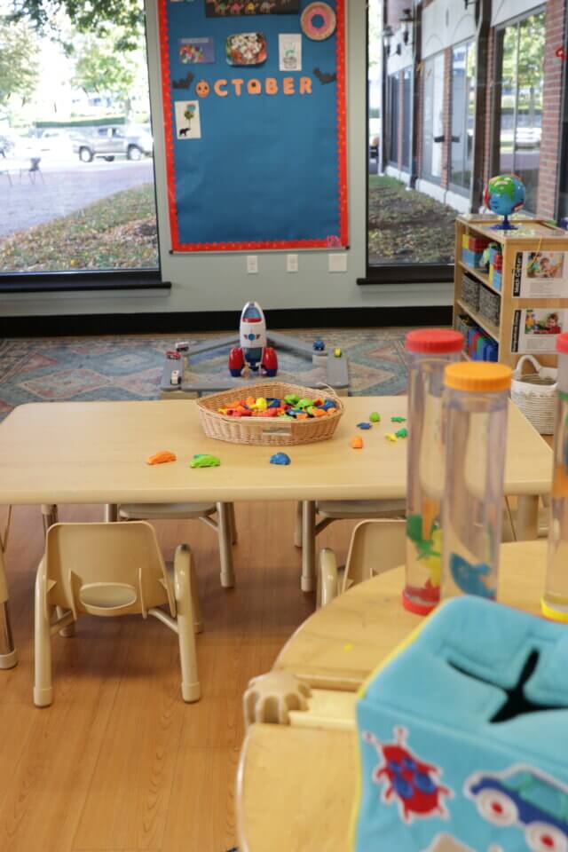 A brightly lit classroom with a small table and chairs, toys scattered on a table, colorful storage bins, and large windows. A bulletin board labeled "October" is on the wall.