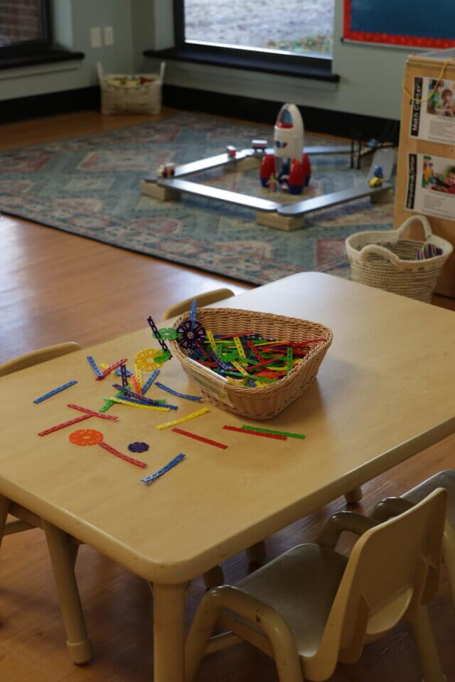 A classroom table with assorted colorful building sticks in a basket and scattered on the surface, children's chairs around the table, and a rug with toys in the background.