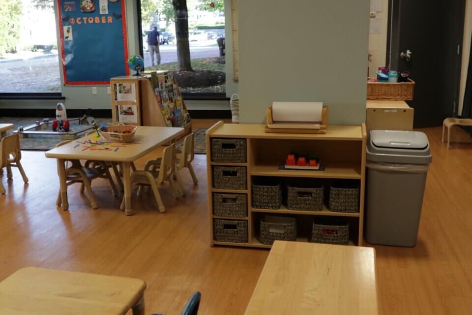 A brightly lit classroom with tables and chairs, storage units with baskets, a bulletin board marked "October," and a large window overlooking trees. A trash can is placed beside a shelving unit.