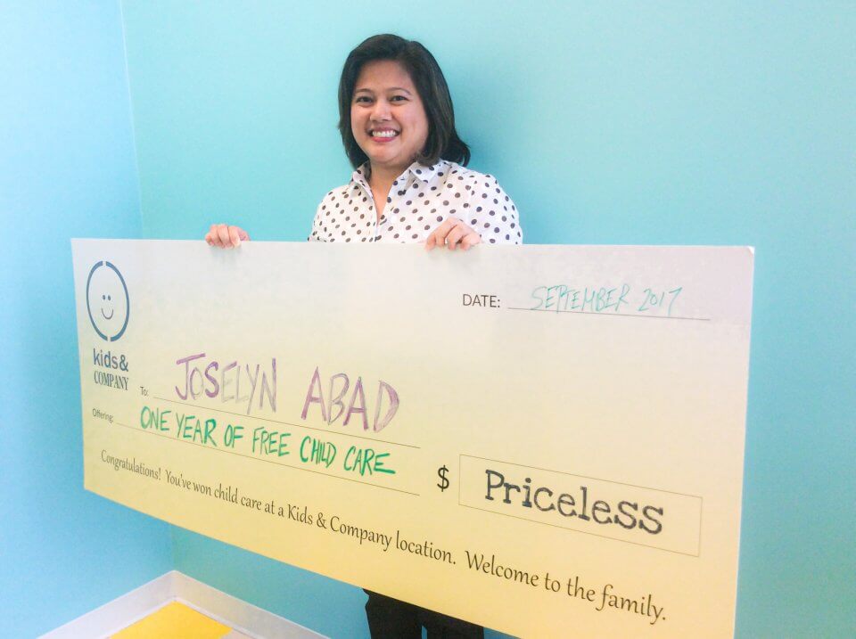 A person smiling and holding a large check for one year of free child care from Kids & Company, dated September 2017. The recipient's name on the check is Josslyn Abad.