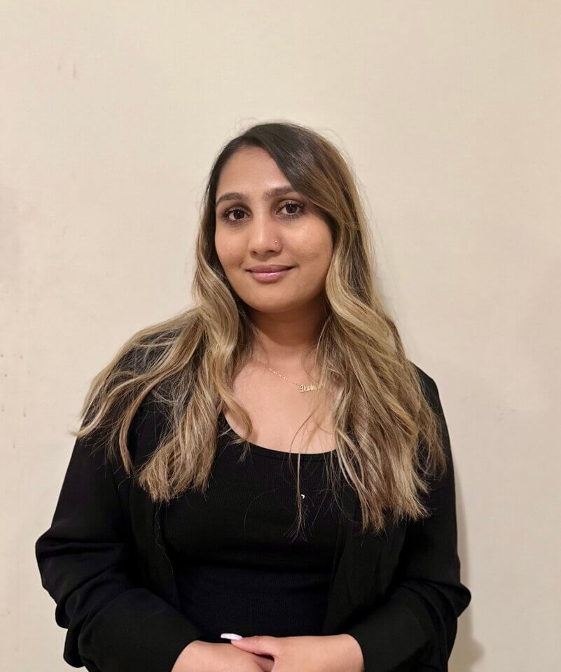 A woman with long wavy hair wearing a black top and black blazer stands against a plain beige wall, looking at the camera.