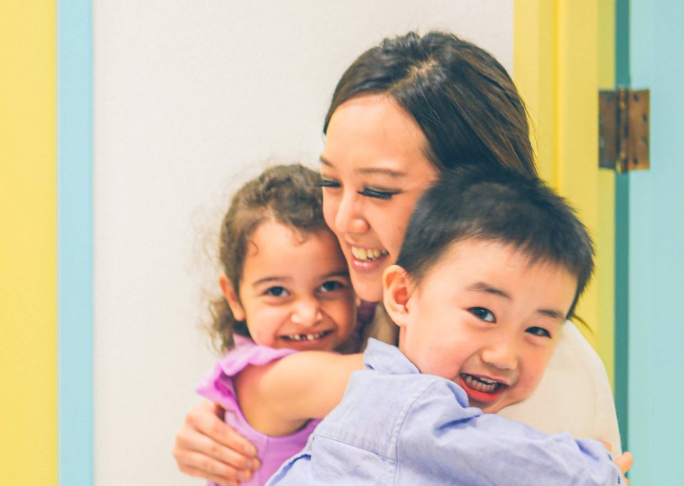 A woman smiles as she embraces two young children, one girl and one boy, in a brightly colored room.