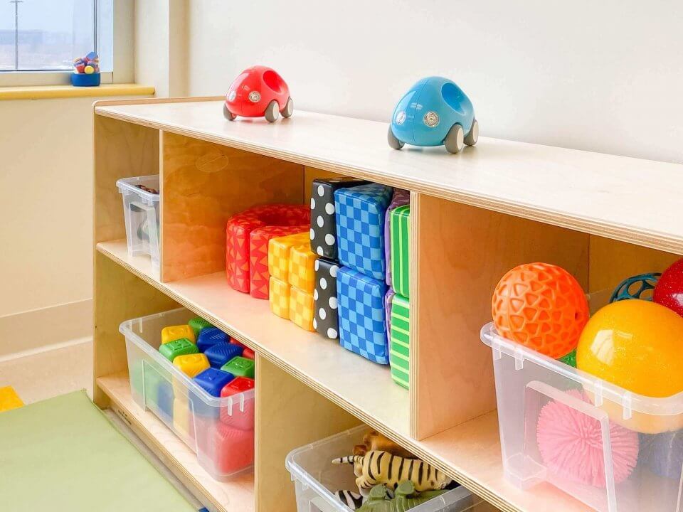 Wooden shelves with toy cars on top and several plastic bins filled with colorful toys, including balls, blocks, and a toy tiger, in an organized playroom.