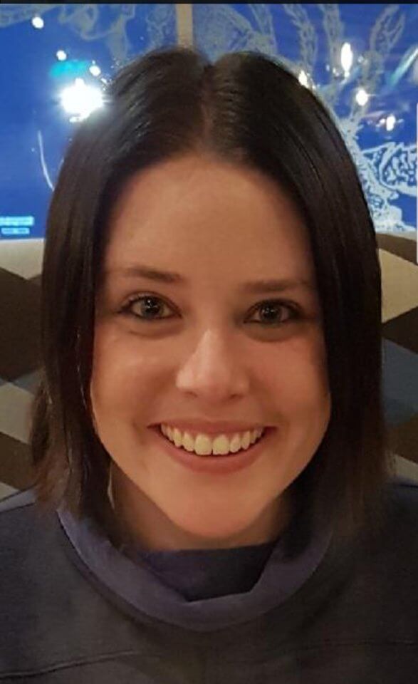 Close-up of a smiling person with straight, shoulder-length dark hair, wearing a dark top, in an indoor setting with a blurred background featuring light reflections.