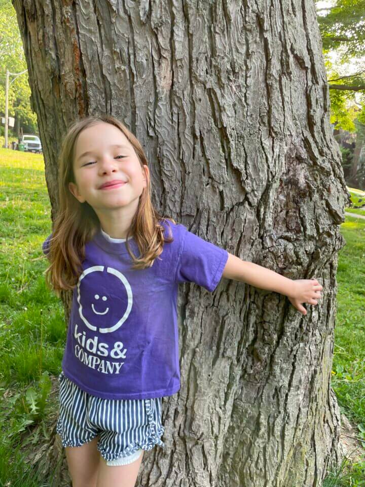 A child wearing a purple "kids & COMPANY" shirt and striped shorts smiles while leaning against a large tree in a grassy area.