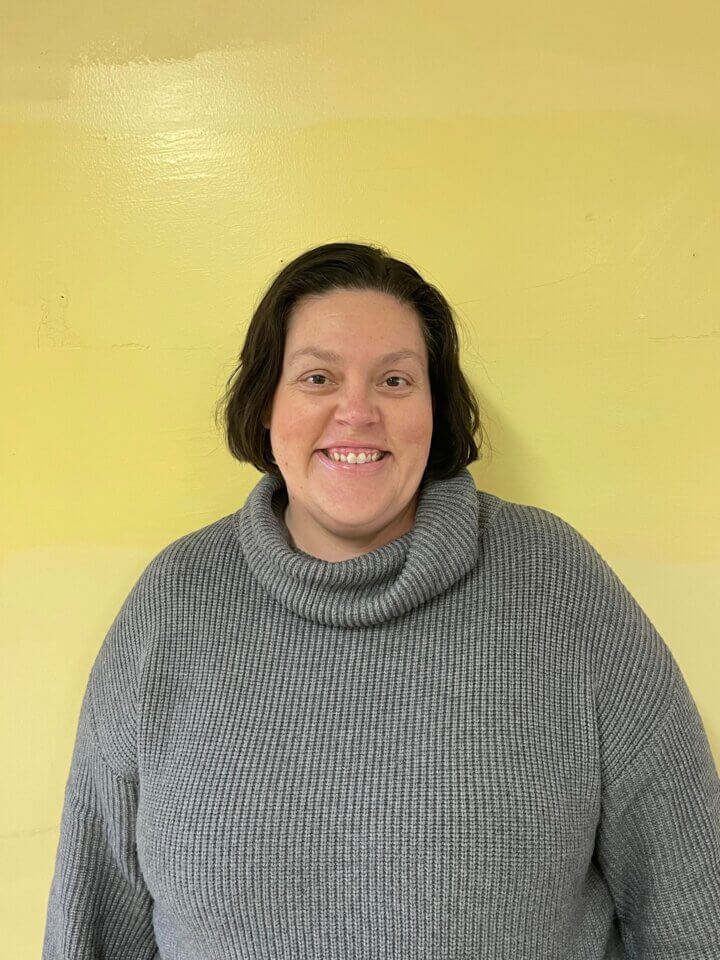 A person with short dark hair wearing a gray turtleneck sweater stands against a yellow wall, smiling at the camera.