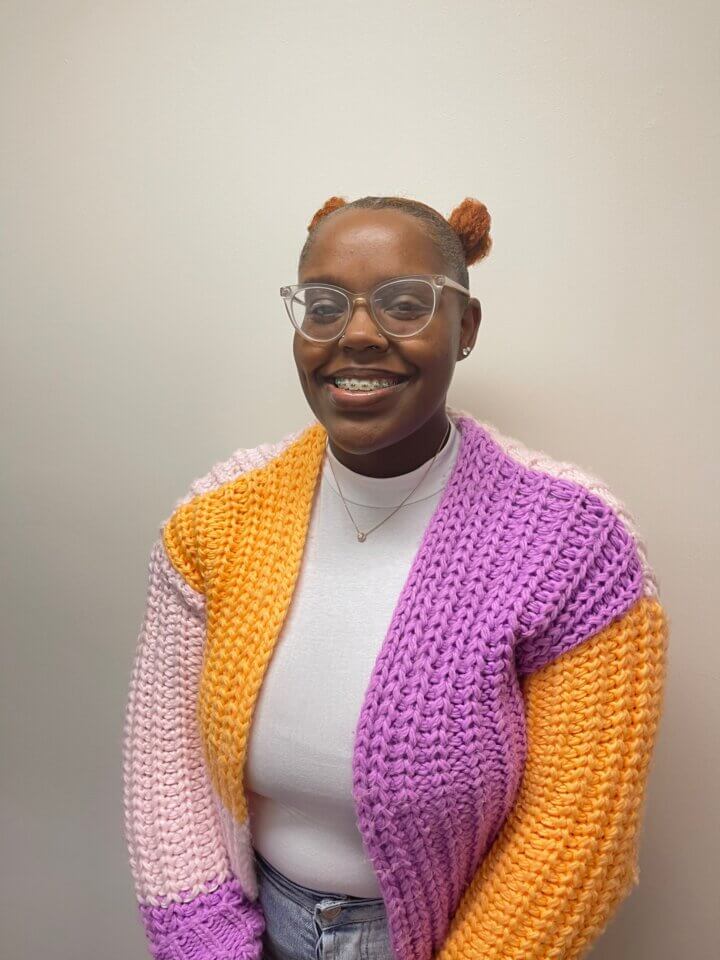 A person wearing glasses, a colorful knitted cardigan, and a white turtleneck smiles while standing against a plain background.