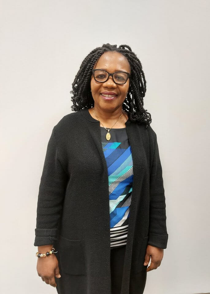 A woman with braided hair, glasses, and a necklace smiles while standing in front of a light-colored background. She is wearing a black cardigan over a patterned top.