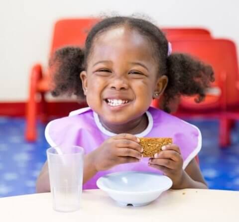 A young girl with a joyful smile, sitting at a table, eating a granola bar and wearing a bib, with a glass of water and bowl nearby.