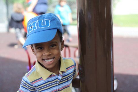 A young child in a blue hat smiling at the camera in a playground setting.