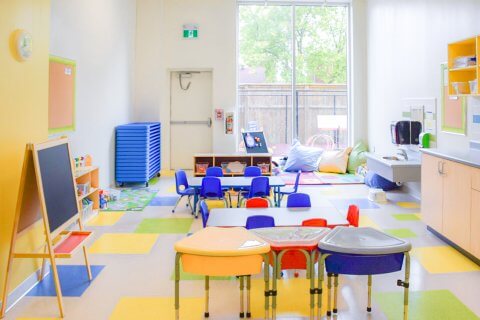 A brightly lit, organized classroom features a small table and chairs, easel, storage shelves, and colorful mats. Large windows and an exit door are situated at the rear.