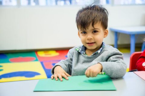 A young boy smiling at the camera while sitting at a table with a green sheet of paper in a colorful classroom.