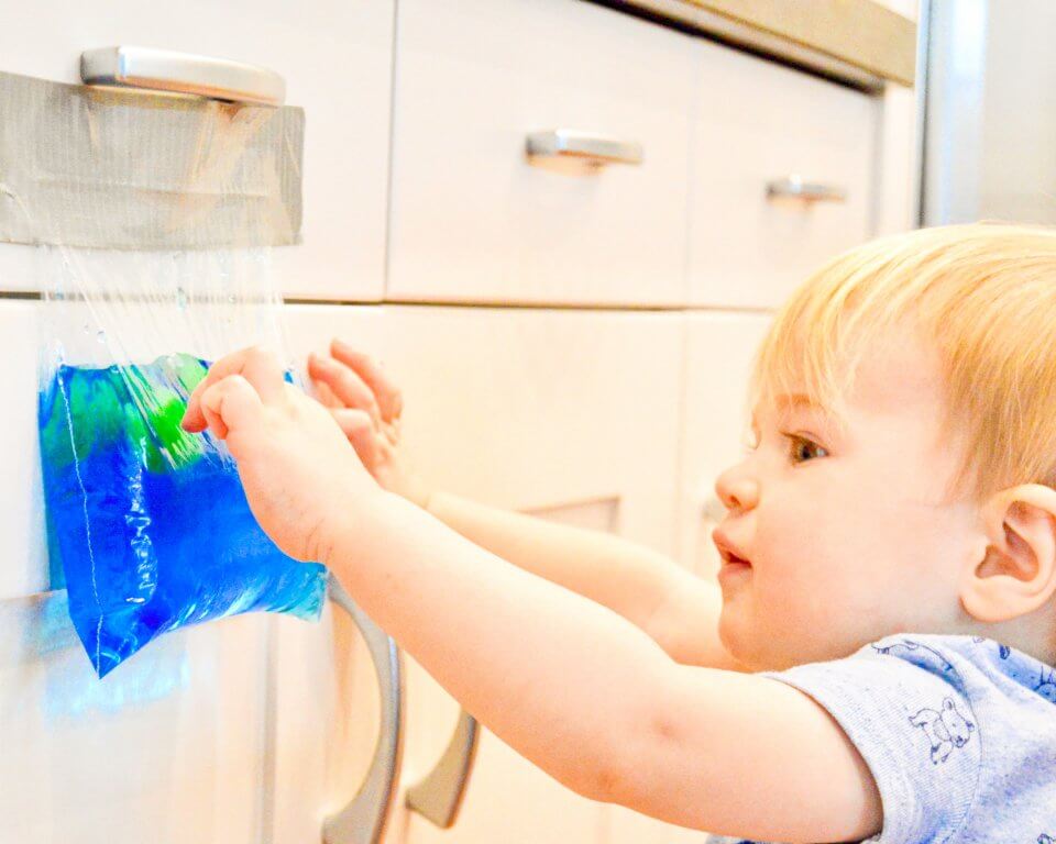 A child is playing with a sensory bag taped filled with hair gel, glitter and food colouring into a secure zipped plastic bag, taped on a kitchen cabinet.