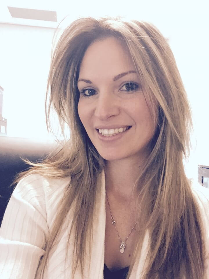 A woman with long, straight hair and a necklace smiles at the camera. She is wearing a light-colored top.