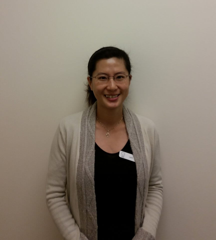 A person with dark hair and glasses stands against a plain wall, wearing a light cardigan over a black top and a name tag.