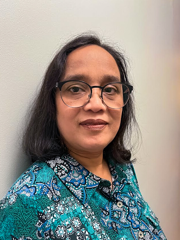 A person with shoulder-length dark hair and glasses wearing a blue patterned shirt stands in front of a plain light-colored wall.