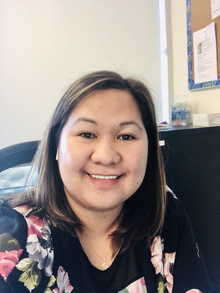 A person with shoulder-length hair wearing a floral-patterned top is smiling at the camera in an office environment. There is a bulletin board and office supplies in the background.